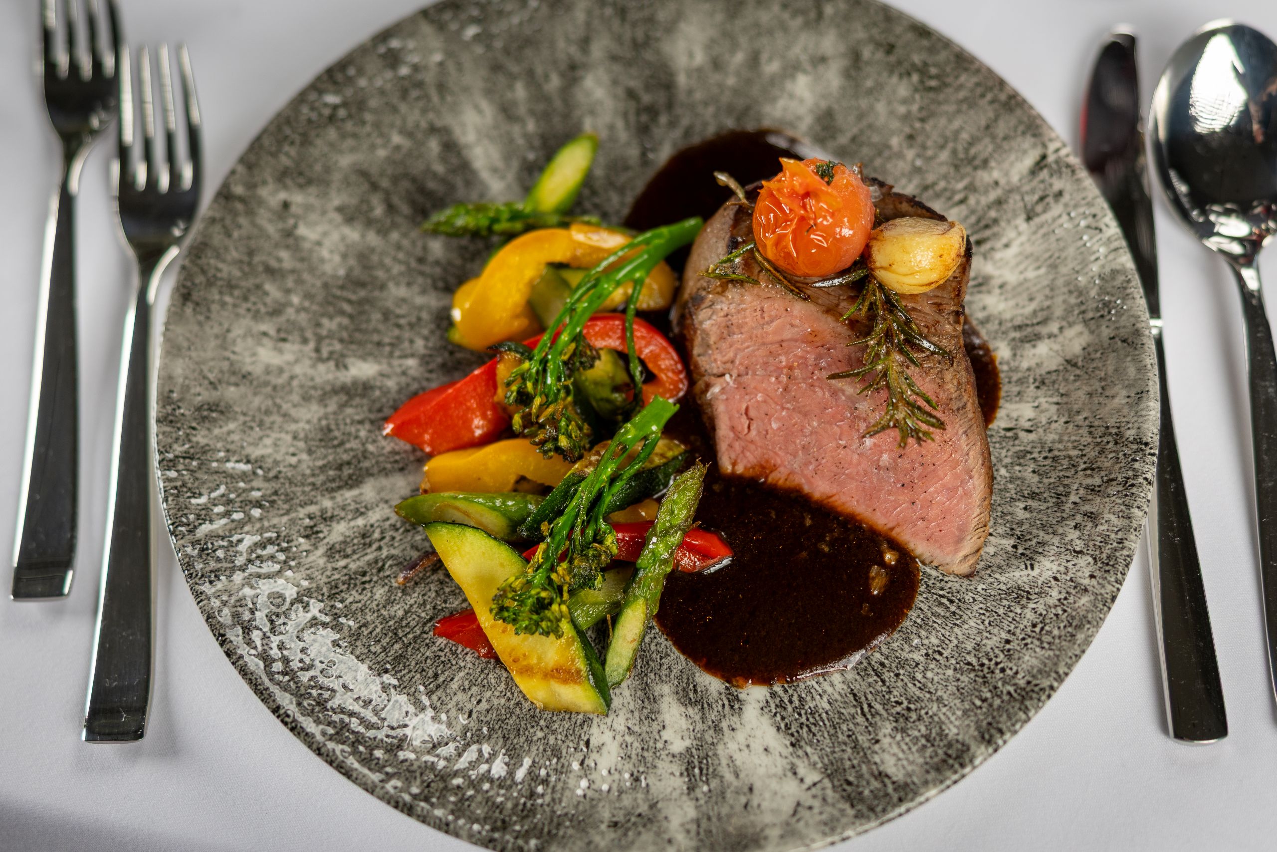 Rosa gebratenes Fleisch mit dunkler Jus und Grillgemüse angerichtet auf einem grauen Teller, daneben Messer und Gabeln sowie ein Löffel.