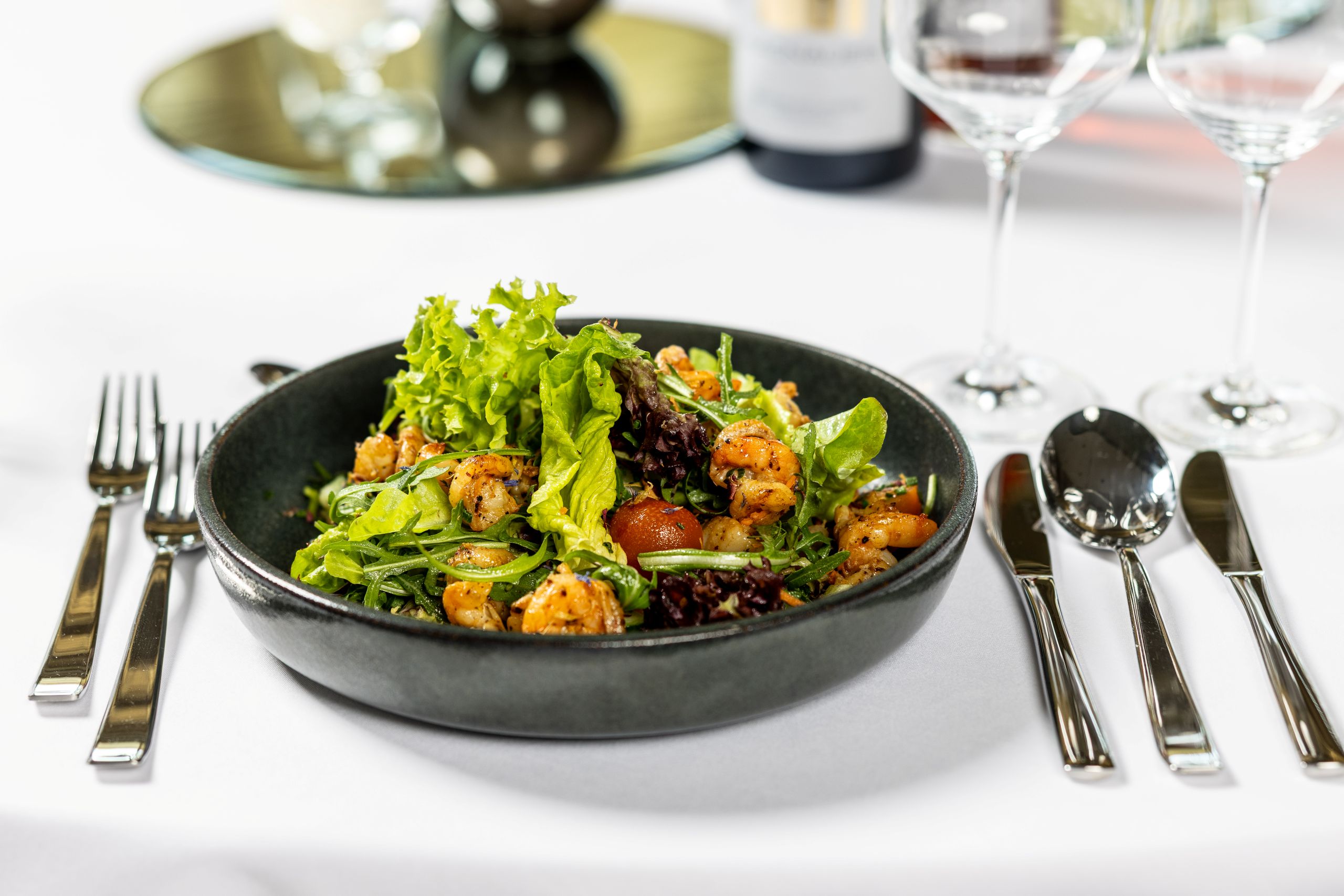 Gemischter grüner Salat mit gebratenen Garnelen in einer schwarzen Schale auf einem eingedeckten Tisch. Links daneben liegen zwei Gabeln, rechts zwei Messer und ein Löffel; verschwommen im Hintergrund stehen zwei Weingläser und eine Weinflasche.