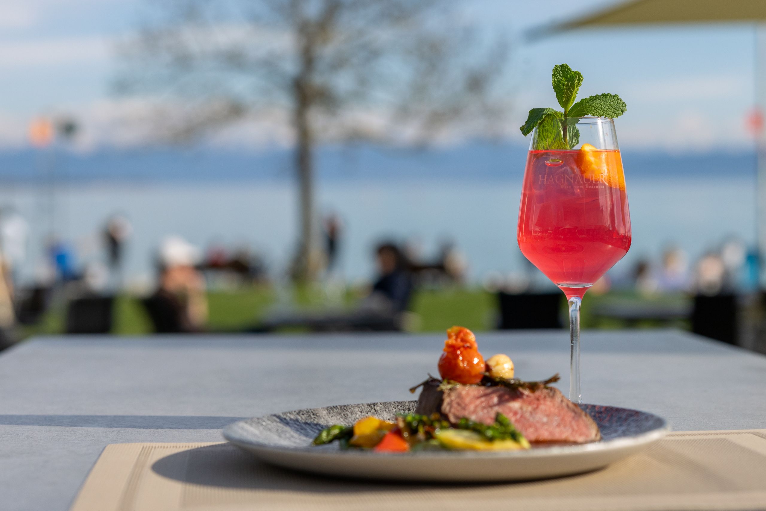 weinglas mit rosafarbenem Getränk dekoriert it Minzezweig und Orangenscheiben steht neben einem dunklen Teller, auf dem gegrilltes Gemüse mit rosa gebratenem Fleisch liegt, auf einem Tisch im Freien. Im Hintergrund verschwommen Menschen und ein See.